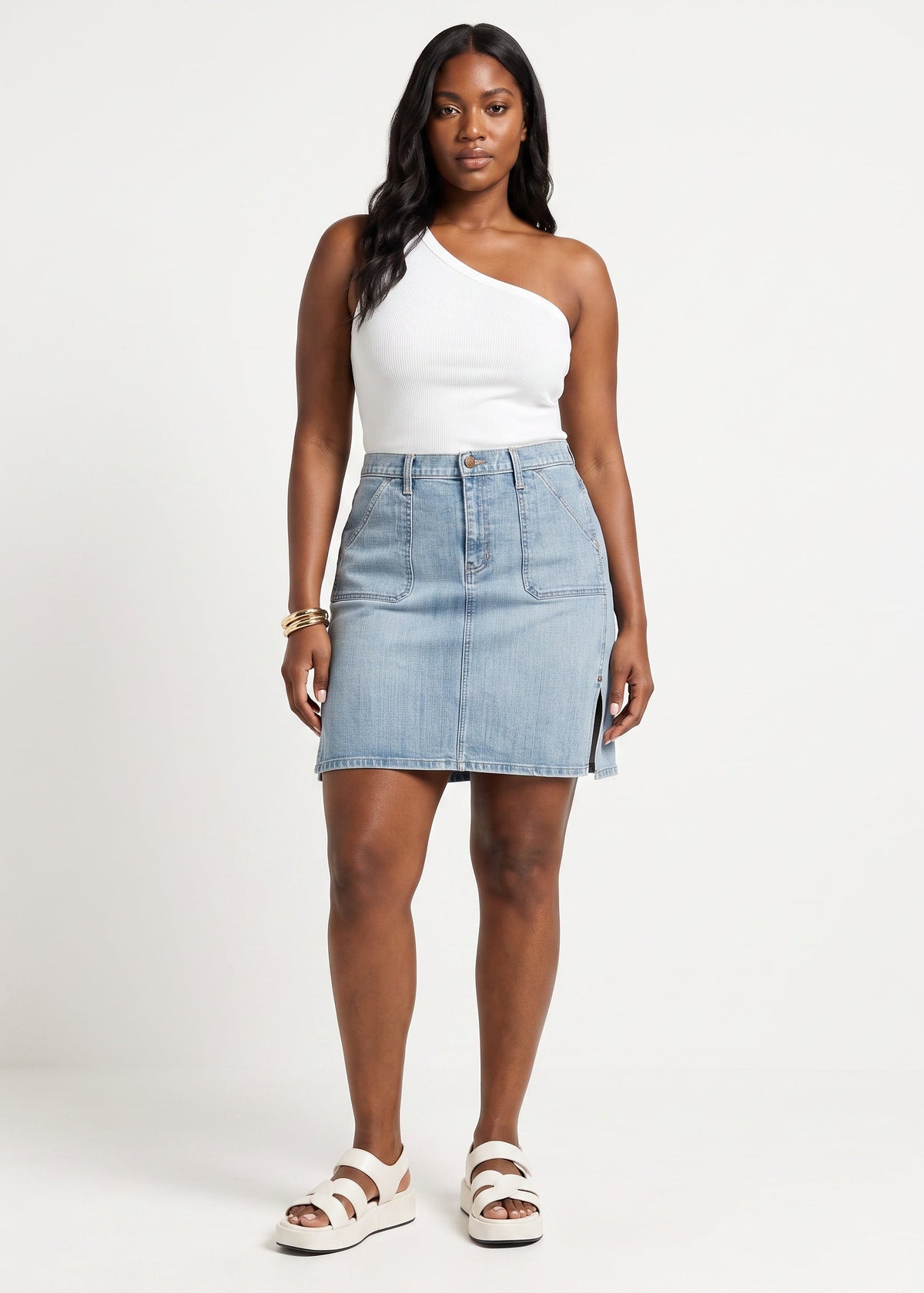 A woman modeling a light-wash denim mini skirt with a side slit, paired with a white one-shoulder top and white slide sandals, against a plain white studio background.