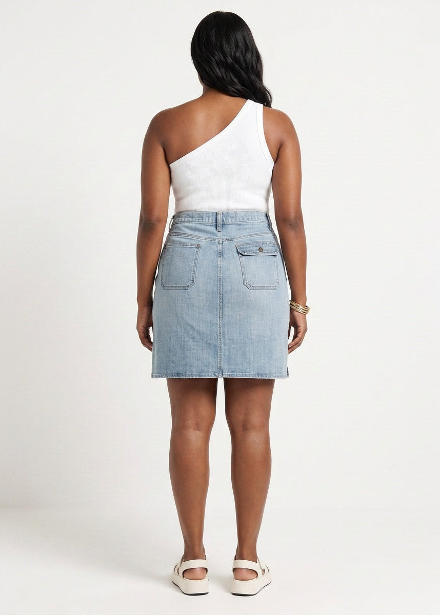 A woman shown from the back wearing a white one-shoulder top and a light blue denim mini skirt with back patch pockets, paired with white slide sandals, against a plain white studio background.