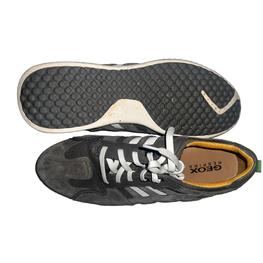 A pair of worn dark gray Geox Respira sneakers, showing the textured black outsole of one shoe and the side profile of the other, with a hand holding the top edge of the sole against a white background with red paint marks.