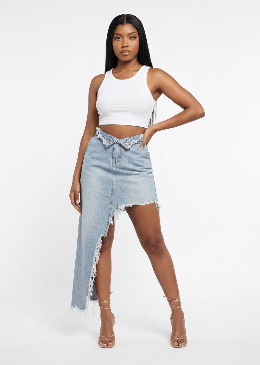 A model wearing a white ribbed crop top and a light-wash denim asymmetrical skirt with fringe and a decorative lace-up detail at the waist, paired with nude high-heeled sandals, against a plain white studio background.