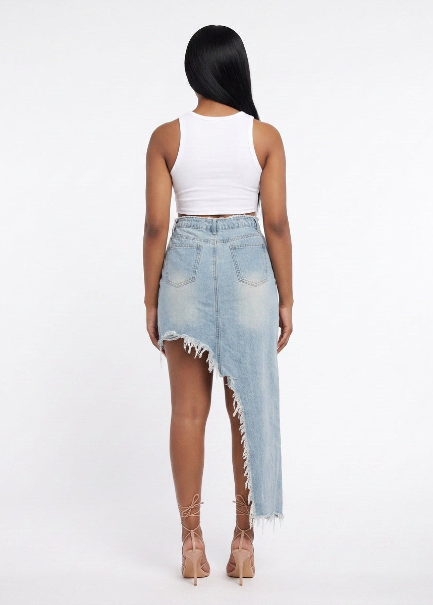 A woman shown from the back wearing a light-wash denim asymmetrical mini skirt with frayed edges, paired with a white ribbed tank top and beige lace-up heels, against a plain white studio background.